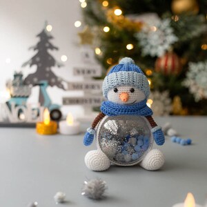 May include: A crocheted snowman with a blue hat, scarf, and mittens, and a carrot nose. The snowman has a clear, round body filled with silver and blue glitter. The snowman is a festive holiday decoration.