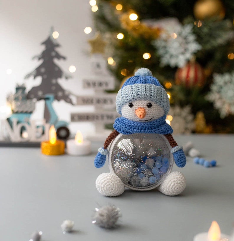 May include: A crocheted snowman with a blue hat, scarf, and mittens, and a carrot nose. The snowman has a clear, round body filled with silver and blue glitter. The snowman is a festive holiday decoration.