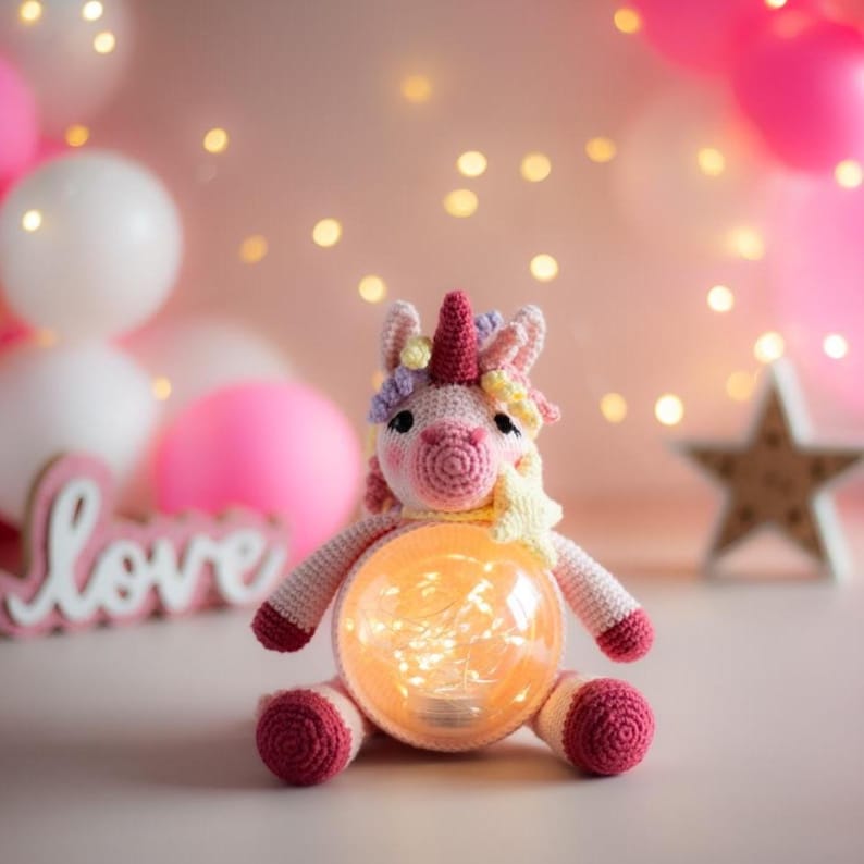 May include: A handmade crochet unicorn lamp with a glowing orb belly. The unicorn is pink, with a red horn, and colorful mane and tail. The lamp is surrounded by pink and white balloons and a wooden star.