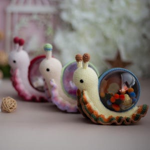 May include: Three crocheted snail toys with colorful shells. The snails are made of yarn and have small black eyes. The shells are filled with colorful beads.