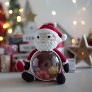 Peut inclure: Un personnage fait &agrave; la main du P&egrave;re No&euml;l au crochet, avec un bonnet rouge, une barbe blanche et des bottes noires. Le P&egrave;re No&euml;l tient un r&eacute;cipient rond transparent rempli de d&eacute;corations roses et dor&eacute;es, parfait pour No&euml;l.