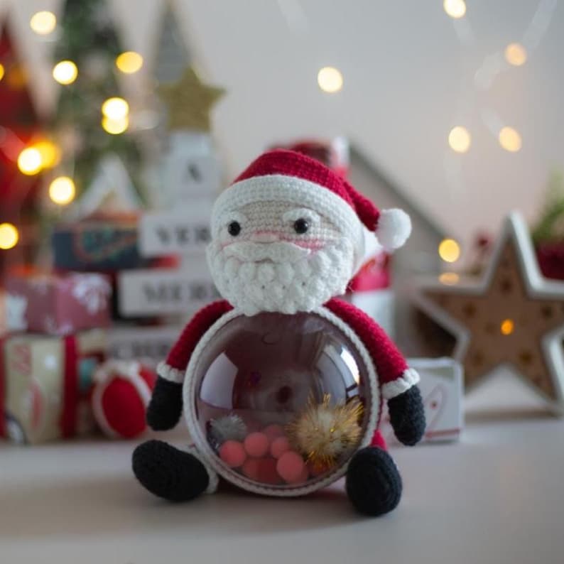 Peut inclure: Un personnage fait &agrave; la main du P&egrave;re No&euml;l au crochet, avec un bonnet rouge, une barbe blanche et des bottes noires. Le P&egrave;re No&euml;l tient un r&eacute;cipient rond transparent rempli de d&eacute;corations roses et dor&eacute;es, parfait pour No&euml;l.
