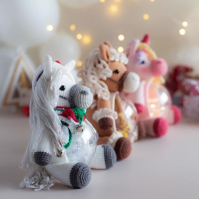 May include: Three handmade crochet unicorn ornaments in a row. The first is white with a red and green scarf and silver bells. The second is brown with a cream mane. The third is pink with a yellow horn. Each is inside a clear glass ball.
