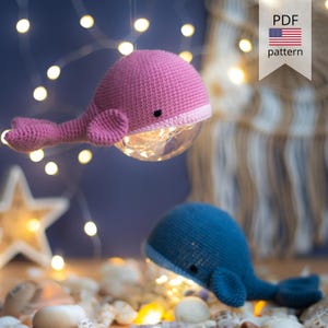 May include: Two crocheted whale-shaped lamps, one pink and one blue, with a clear globe belly containing fairy lights. The pink whale is suspended from above. The lamps are surrounded by seashells and a string of lights. A banner with "PDF pattern" is visible.