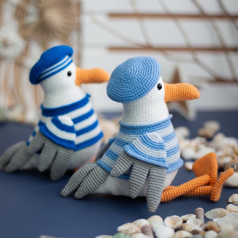 May include: Two handmade crochet seagull plush toys. Each seagull is white with grey wings and feet, an orange beak, and a blue hat. One wears a blue and white striped shirt, the other a blue beret. The toys are on a blue surface.