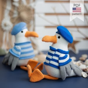 May include: Two handmade crochet seagull toys. Each seagull wears a blue beret and a blue and white striped shirt. The seagulls have orange beaks and feet, and grey wings. The image includes a banner that says "PDF pattern".