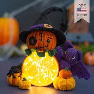 May include: Crocheted Halloween pumpkin wearing a witch's hat, with a crooked smile and a single eye. The pumpkin is holding a string of lights and is surrounded by smaller pumpkins and a purple crocheted bat.