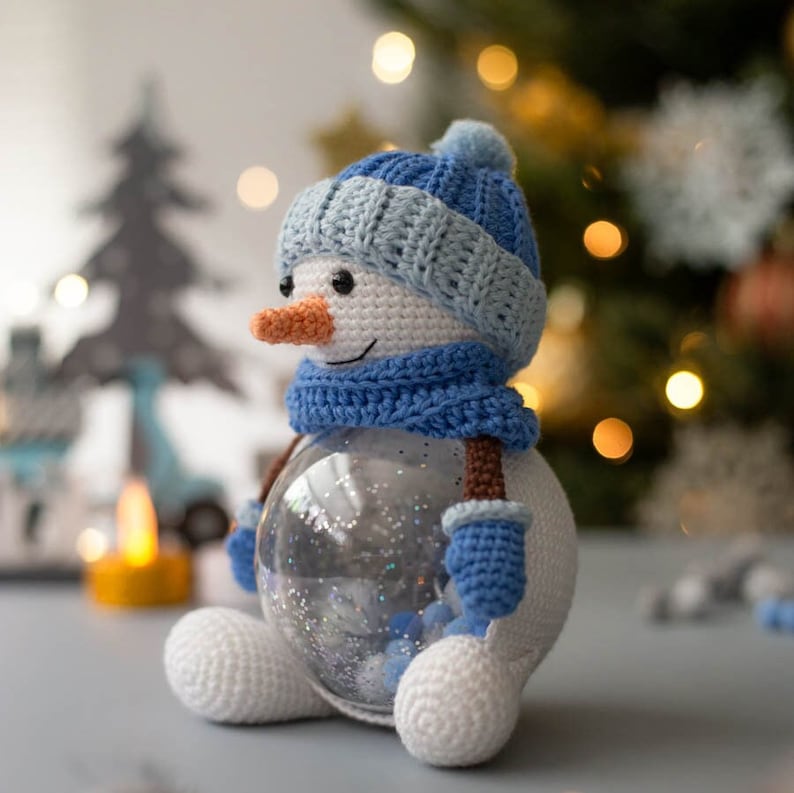 May include: A crocheted snowman with a blue hat, scarf, and mittens. The snowman has a carrot nose and a clear glass globe body filled with glitter. The snowman is sitting on a gray surface, with a blurred background of a Christmas tree.