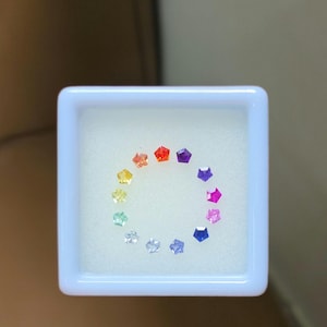 Puede incluir: Una colección de gemas coloridas en forma de estrella dispuestas en círculo dentro de un recipiente cuadrado blanco. Las gemas muestran un espectro de colores, incluyendo rojo, naranja, amarillo, verde, azul y morado.