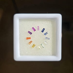 Può includere: Una scatola quadrata bianca contiene una disposizione circolare di pietre preziose rettangolari di vari colori. Le pietre preziose includono sfumature di viola, rosa, blu, verde, giallo, arancione e trasparente. Le pietre sono su una superficie bianca strutturata.
