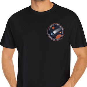 May include: Black t-shirt featuring a circular graphic with a comet, planets, and text. The design is in shades of orange, blue, and white. The shirt is made of a soft material and is being worn.