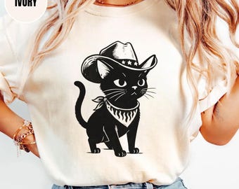 Op de afbeelding: Ivoor kleurig t-shirt met een zwarte cartoonkat met een cowboyhoed en bandana. De kat heeft een gedetailleerd ontwerp met snorharen en een gebogen staart. Het woord "IVORY" staat in een witte cirkel linksboven. Het shirt wordt door een persoon gedragen.