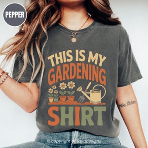 May include: Dark grey t-shirt with the text "THIS IS MY GARDENING SHIRT" in orange, yellow, and green. The design features potted flowers, a watering can, and gardening tools. The shirt is a comfortable, casual style.