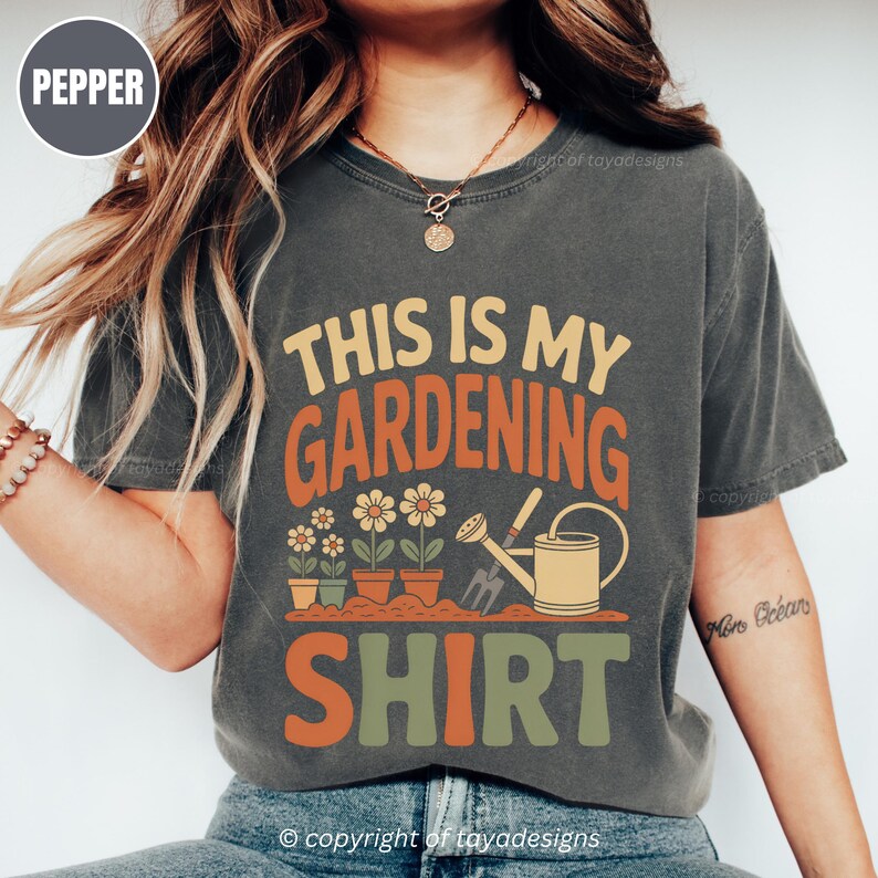 May include: Dark grey t-shirt with the text "THIS IS MY GARDENING SHIRT" in orange, yellow, and green. The design features potted flowers, a watering can, and gardening tools. The shirt is a comfortable, casual style.