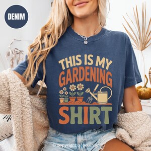 May include: Denim blue t-shirt with the text "THIS IS MY GARDENING SHIRT" in orange, green, and cream colours. The shirt features a graphic of potted flowers, a watering can, and a gardening tool. The shirt is a casual style.