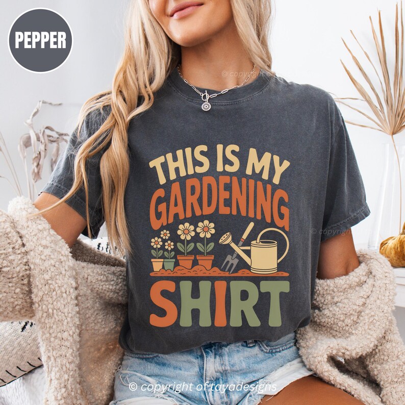 May include: Dark grey t-shirt with the text "THIS IS MY GARDENING SHIRT" in orange, yellow, and green. The shirt features a graphic of flowers, pots, a watering can, and a gardening tool.
