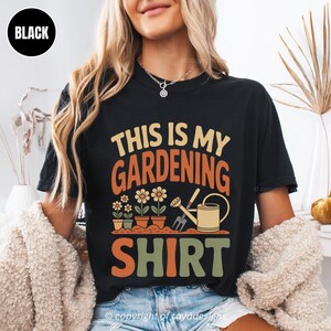 May include: Black t-shirt featuring the text "THIS IS MY GARDENING SHIRT" in orange, green, and tan. The design includes illustrations of flowers in pots, a watering can, and a gardening fork. A casual, comfortable style.
