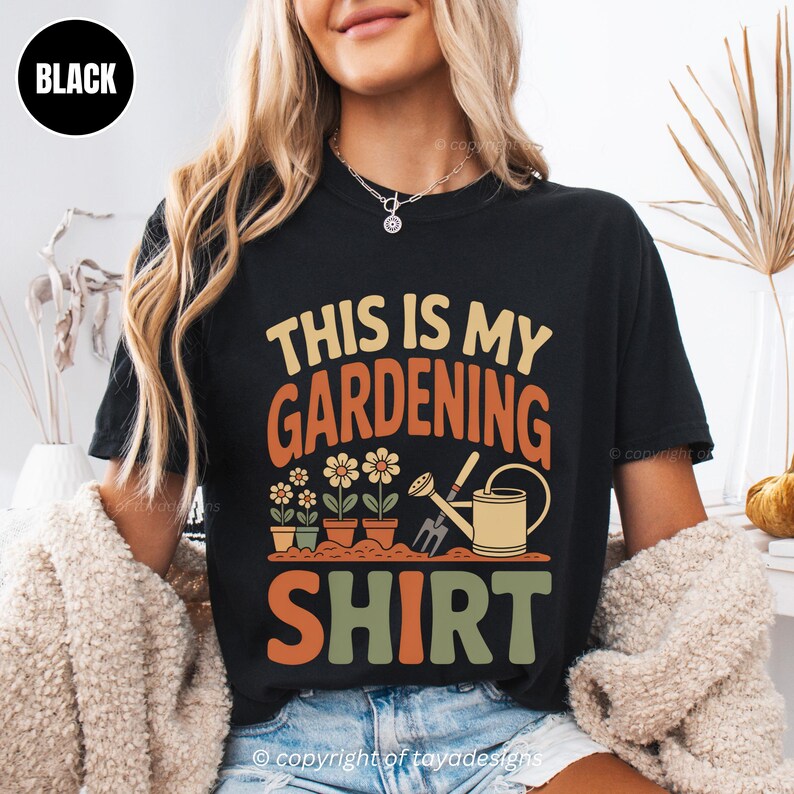 May include: Black t-shirt featuring the text "THIS IS MY GARDENING SHIRT" in orange, green, and tan. The design includes illustrations of flowers in pots, a watering can, and a gardening fork. A casual, comfortable style.