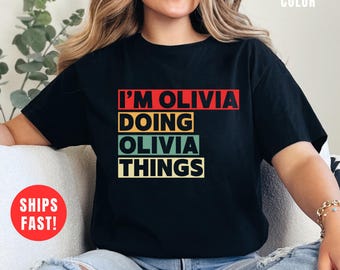 May include: Black t-shirt with the text "I'M OLIVIA DOING OLIVIA THINGS" in red, yellow, green, and white block letters. The shirt is a crew neck style. The words "BLACK COLOR" are in the upper right corner.