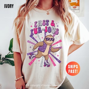May include: Ivory-colored t-shirt featuring a cartoon sloth wearing sunglasses and roller skates, with the text "Fast & Fur-ious" in purple and pink. The sloth is set against a backdrop of radiating purple and pink lines.