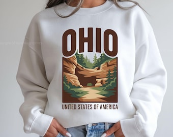 Könnte beinhalten: Weißes Sweatshirt mit brauner Grafik des Wortes "OHIO" über einer Landschaftsszene mit Weg, Bäumen und Felsformationen. Der Text "UNITED STATES OF AMERICA" steht darunter. Das Sweatshirt hat lange Ärmel und einen Rundhalsausschnitt.
