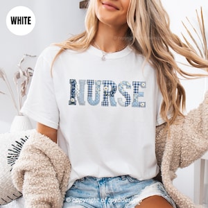 Puede incluir: Camiseta blanca con la palabra "NURSE" en letras azules de patchwork. Las letras presentan patrones florales, de cuadros y de lunares. La camiseta es de cuello redondo y tiene mangas cortas.