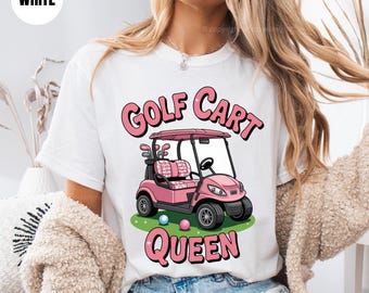 May include: White t-shirt with a pink golf cart graphic and the text "Golf Cart Queen." The golf cart is pink with golf clubs in the back. Two golf balls are on the green grass.