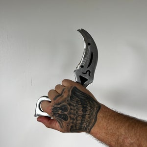 May include: A silver metal karambit knife with a ring handle. The knife is held in a hand with a skull tattoo.