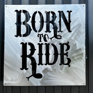 May include: A silver metal sign with black lettering that reads "Born to Ride". The letters are in a stylized font.