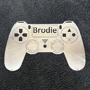 May include: A silver metal cutout of a video game controller with the name "Brodie" in the center.