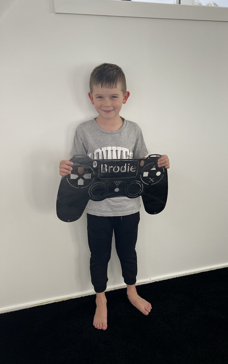 May include: A young boy is holding a black metal cutout of a video game controller. The controller has the name "Brodie" written on it.