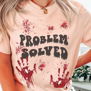 Può includere: Una maglietta color pesca con il testo "PROBLEM SOLVED" in lettere nere che gocciolano. La maglietta ha impronte di mani rosse e schizzi di sangue.
