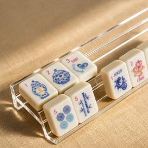 Può includere: Un supporto in acrilico trasparente che contiene diverse tessere del Mahjong color bianco sporco. Le tessere presentano disegni stampati in blu e rosa, tra cui un vaso per lo zenzero, una farfalla, un pesce con l'etichetta JOKER, un drago con l'etichetta CRAK, steli di bambù e motivi floreali.