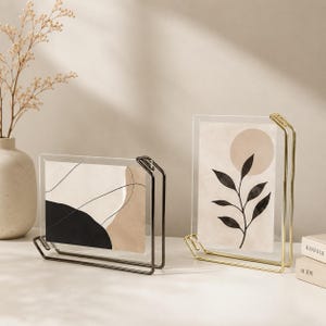 May include: Two rectangular acrylic photo frames with metal stands. One frame is horizontal with a black and beige abstract print, featuring a dark metal stand. The other is vertical with a beige and black botanical print, featuring a gold metal stand.