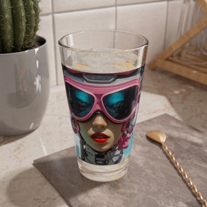 May include: A clear glass with a colorful image of a woman wearing pink ski goggles and a futuristic helmet. The glass is filled with a clear liquid and ice.