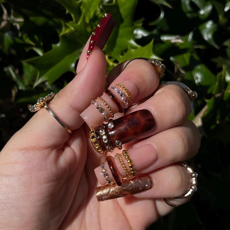 Op de afbeelding: Een hand met lange, verzorgde nagels en meerdere ringen. De nagels hebben verschillende ontwerpen, waaronder effen kleuren, gouden accenten en een schildpadpatroon. De ringen zijn goud en zilver, sommige met edelstenen.