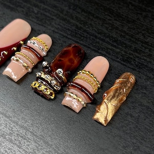 Op de afbeelding: Close-up van verschillende lange, kunstmatige nagels in verschillende ontwerpen. E&eacute;n nagel is nude van kleur met een donkerrode punt. Andere nagels hebben gouden en zilveren ketting- en kraalaccenten en een schildpadontwerp. E&eacute;n nagel is goud.
