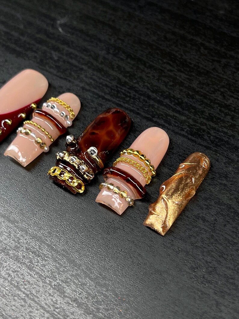 Op de afbeelding: Close-up van verschillende lange, kunstmatige nagels in verschillende ontwerpen. E&eacute;n nagel is nude van kleur met een donkerrode punt. Andere nagels hebben gouden en zilveren ketting- en kraalaccenten en een schildpadontwerp. E&eacute;n nagel is goud.