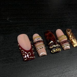Op de afbeelding: Een verzameling van zes kunstnagels. De nagels hebben verschillende ontwerpen, waaronder een nude basis met een bordeauxrode French tip en gouden versieringen, en nagels met gouden en zilveren kettingaccenten. Andere nagels hebben een schildpadpatroon.