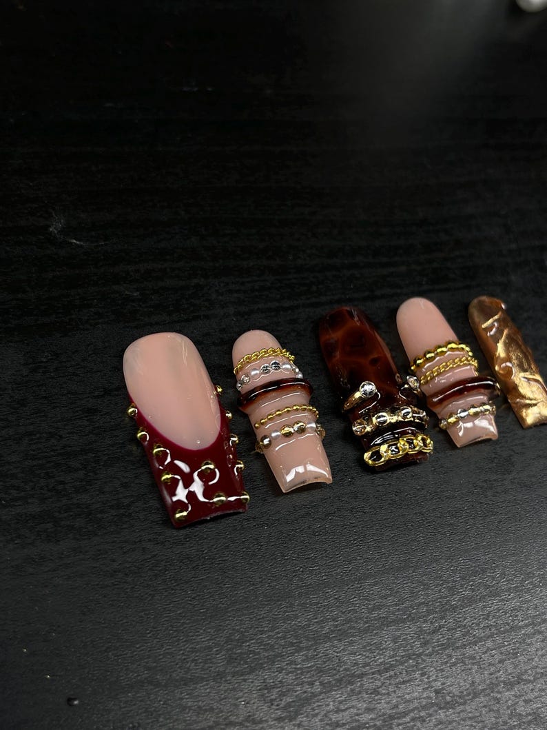 Op de afbeelding: Een verzameling van zes kunstnagels. De nagels hebben verschillende ontwerpen, waaronder een nude basis met een bordeauxrode French tip en gouden versieringen, en nagels met gouden en zilveren kettingaccenten. Andere nagels hebben een schildpadpatroon.