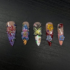 Può includere: Cinque unghie finte lunghe e appuntite con diversi disegni di farfalle e fiori. Le unghie sono dipinte in vari colori, tra cui rosa, bianco, giallo, blu e rosso. Ogni unghia è decorata con strass.