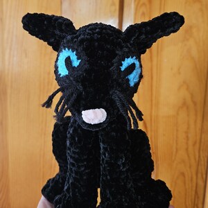 May include: A black crocheted cat plush toy with blue eyes and a pink nose. The cat is sitting on a hand.