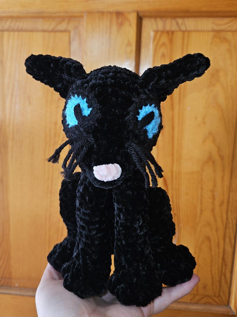 May include: A black crocheted cat plush toy with blue eyes and a pink nose. The cat is sitting on a hand.