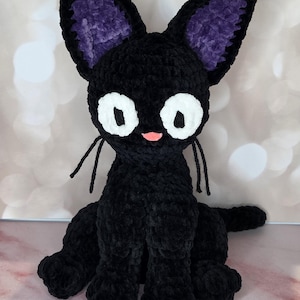 May include: A handmade crochet plush cat made of black yarn with large white circular eyes, a small pink nose, and long black yarn whiskers. The cat features tall pointed ears with purple inner sections and a soft, textured velvet-like appearance.