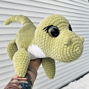 May include: A light green crocheted dinosaur plush toy with large, glittery eyes and a white belly. The handmade stuffed animal has a textured appearance and is designed to resemble a friendly dinosaur.