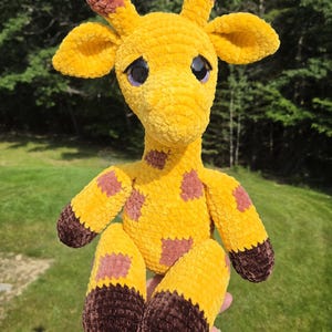 May include: A plush giraffe toy crafted from yellow yarn with a soft, velvety texture. The giraffe features brown patches on its body, dark brown hooves, and two small horns on its head. It has large, round, black plastic eyes with purple accents.