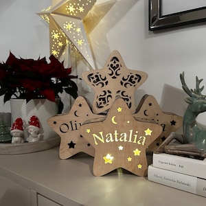 Puede incluir: Tres luces nocturnas de madera en forma de estrella con los nombres Olivia, Natalia y un tercer nombre que está parcialmente oculto. Las estrellas están iluminadas con luces LED blancas cálidas y están dispuestas sobre una mesa con una planta de poinsettia en maceta, dos pequeñas figuras y una pila de libros.
