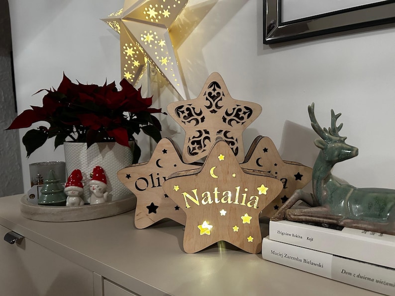 May include: Wooden star-shaped decorative lights with cut-out designs and the name "Natalia" illuminated. A large star-shaped light is in the background. A green deer statue and a poinsettia plant are also visible.