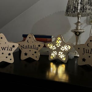 May include: Wooden star-shaped night lights with personalized names: Oliwia, Natalia, and Julia. Each star has a crescent moon and star cutouts. One star has an intricate design. The lights are on a dark surface.