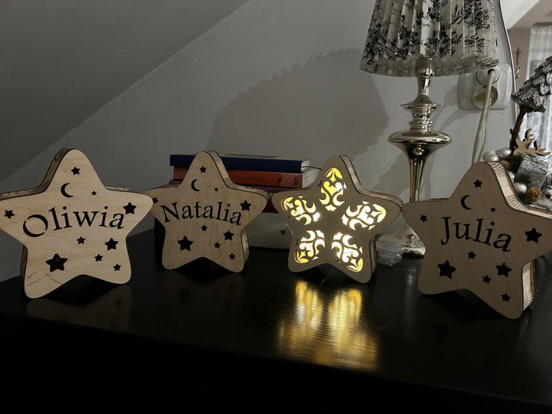 May include: Wooden star-shaped night lights with personalized names: Oliwia, Natalia, and Julia. Each star has a crescent moon and star cutouts. One star has an intricate design. The lights are on a dark surface.